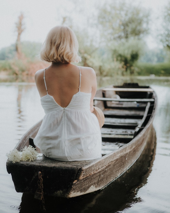 Blonďatá mladá žena v bílých šatech sedí v rybářské lodi