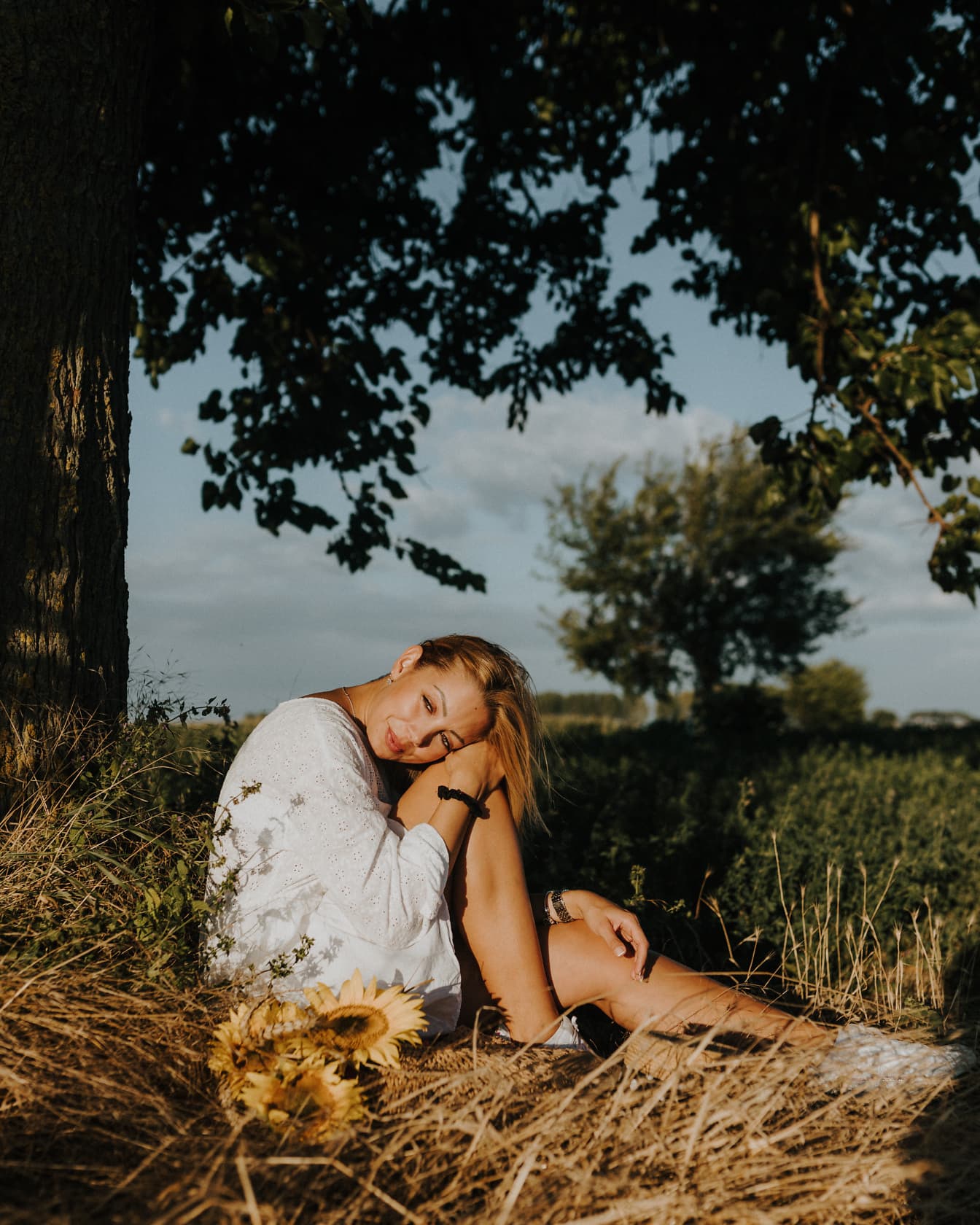 Nádherná blondýnka sedí na venkovské louce a opaluje se při západu slunce