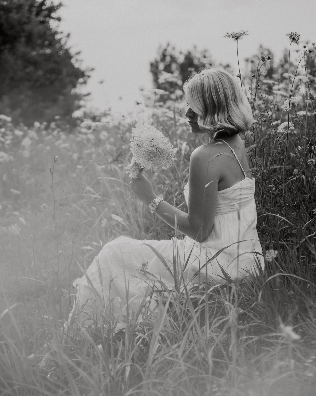 牧草地に座っている白い花のブーケットを持つ白いドレスの女性のモノクロの肖像画