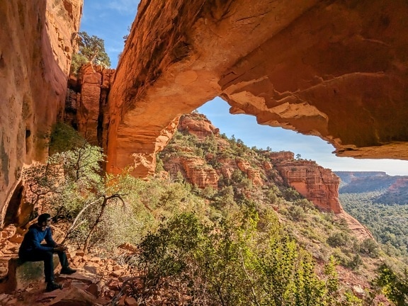 Hiker sitting in shadow of sandstone arch rock formation and enjoying desert panorama