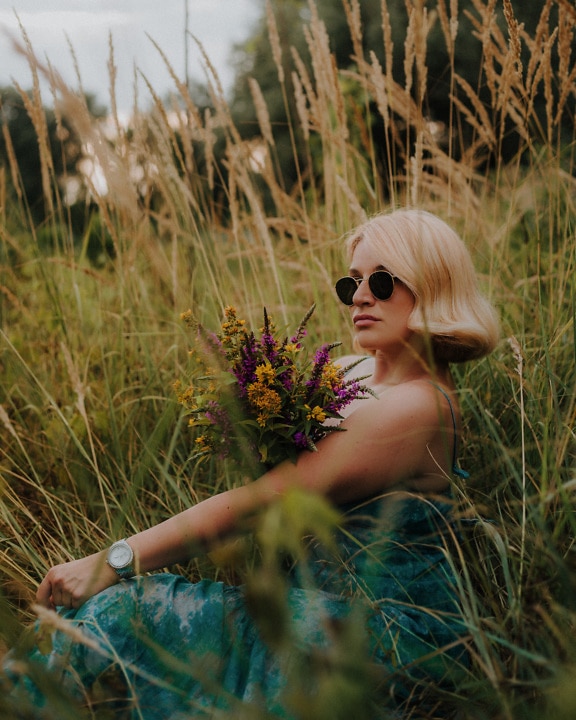 Modelo de foto de mujer joven rubia glamorosa posando en plantas de hierba con ramo de flores silvestres
