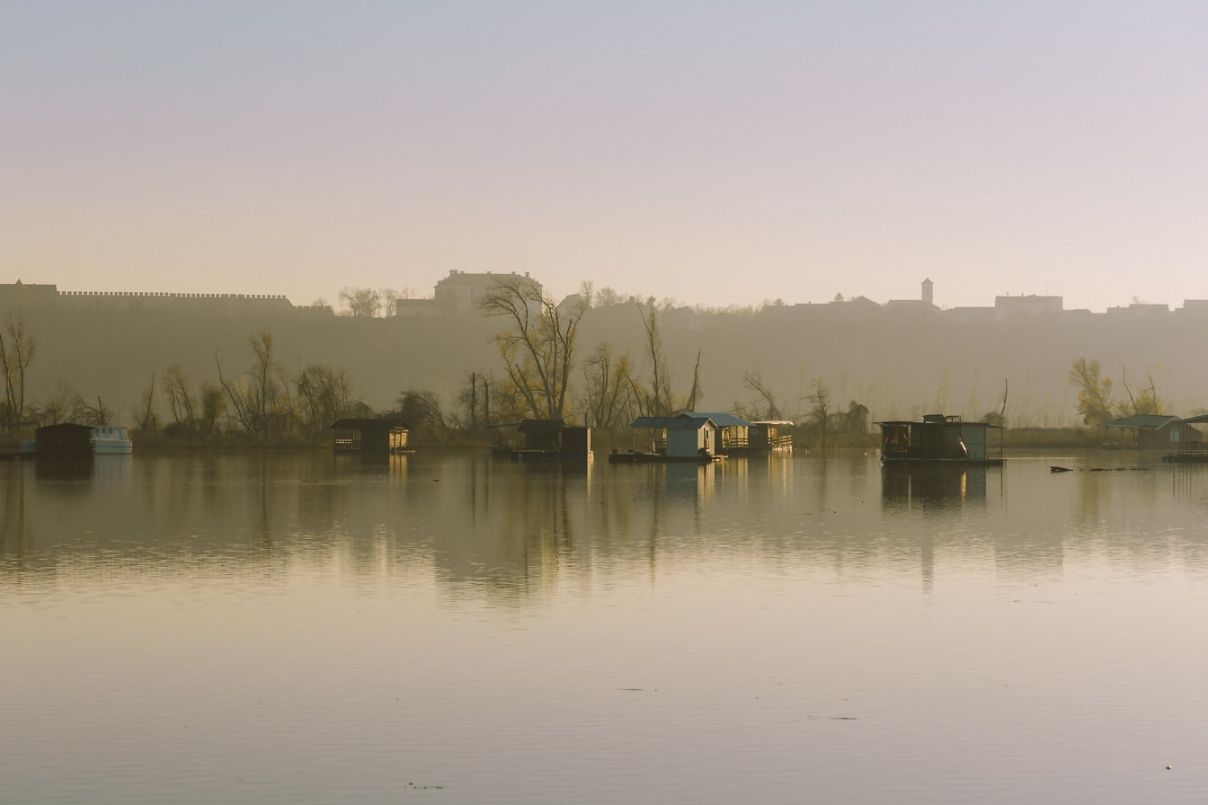 Matahari terbit berkabut di tepi danau dengan rumah perahu di atas air yang tenang