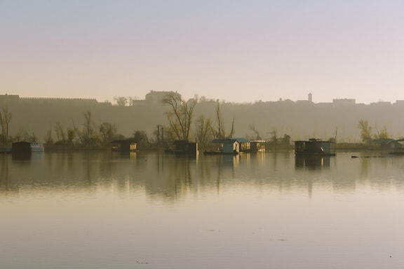 Matahari terbit berkabut di tepi danau dengan rumah perahu di atas air yang tenang