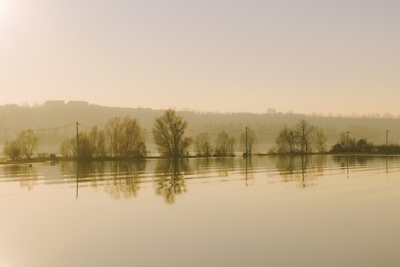 Mlhavé ráno na břehu jezera se siluetou stráně v pozadí