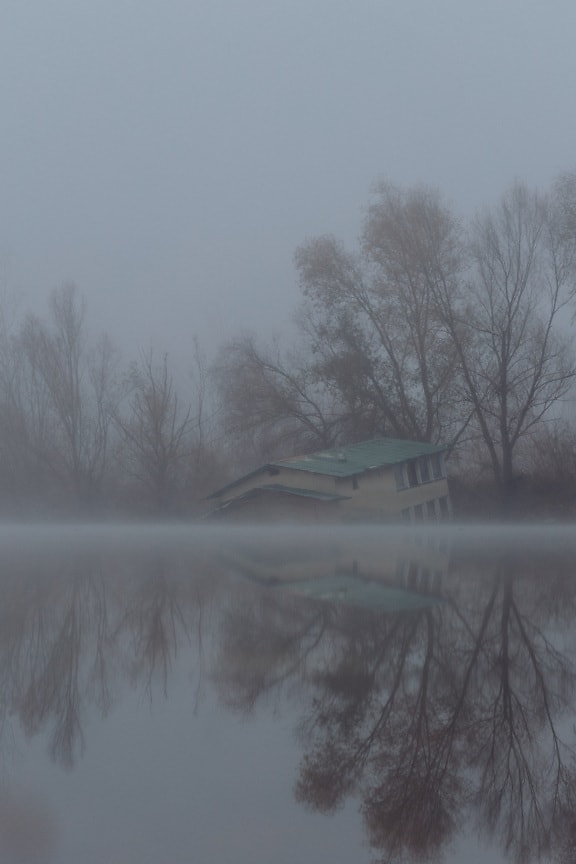 Flooded boathouse on riverbank of foggy lakeside