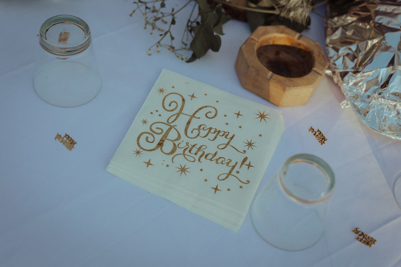 Servilleta de papel blanco con texto de brillo dorado de feliz cumpleaños en la mesa