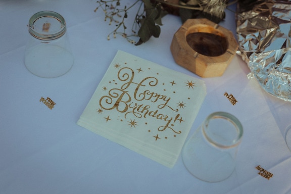 Obrúsok z bieleho papiera s textom Všetko najlepšie k narodeninám so zlatým leskom na stole