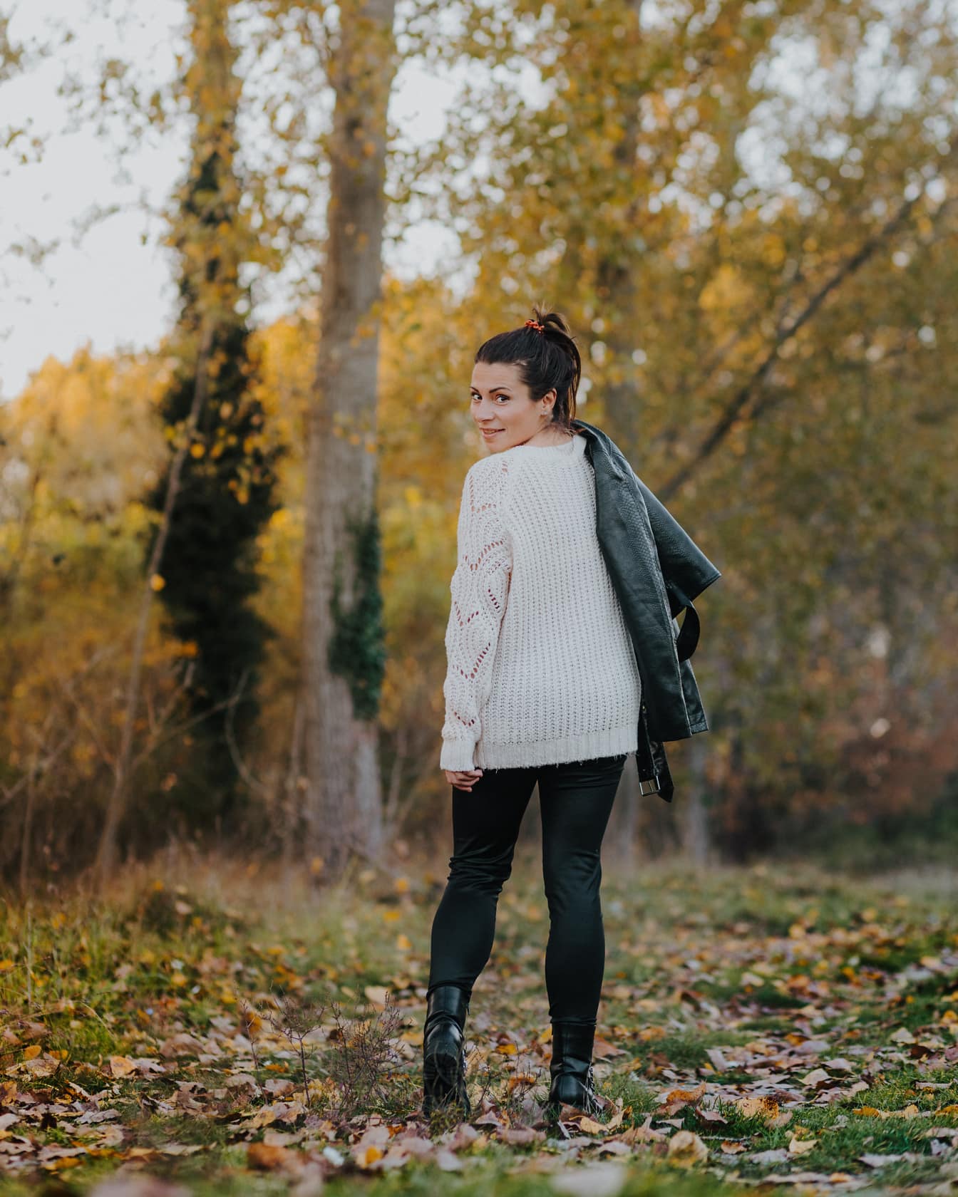 Woman in white cardigan and black leather jacket and boots in forest
