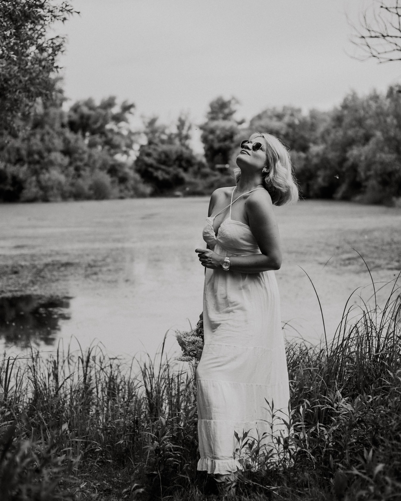 Monokromt portrett av glamourblond ung kvinne i hvit kjole ved innsjøen