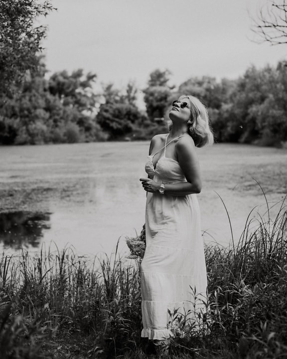 Retrato monocromo de una joven rubia glamurosa en vestido blanco en la orilla del lago