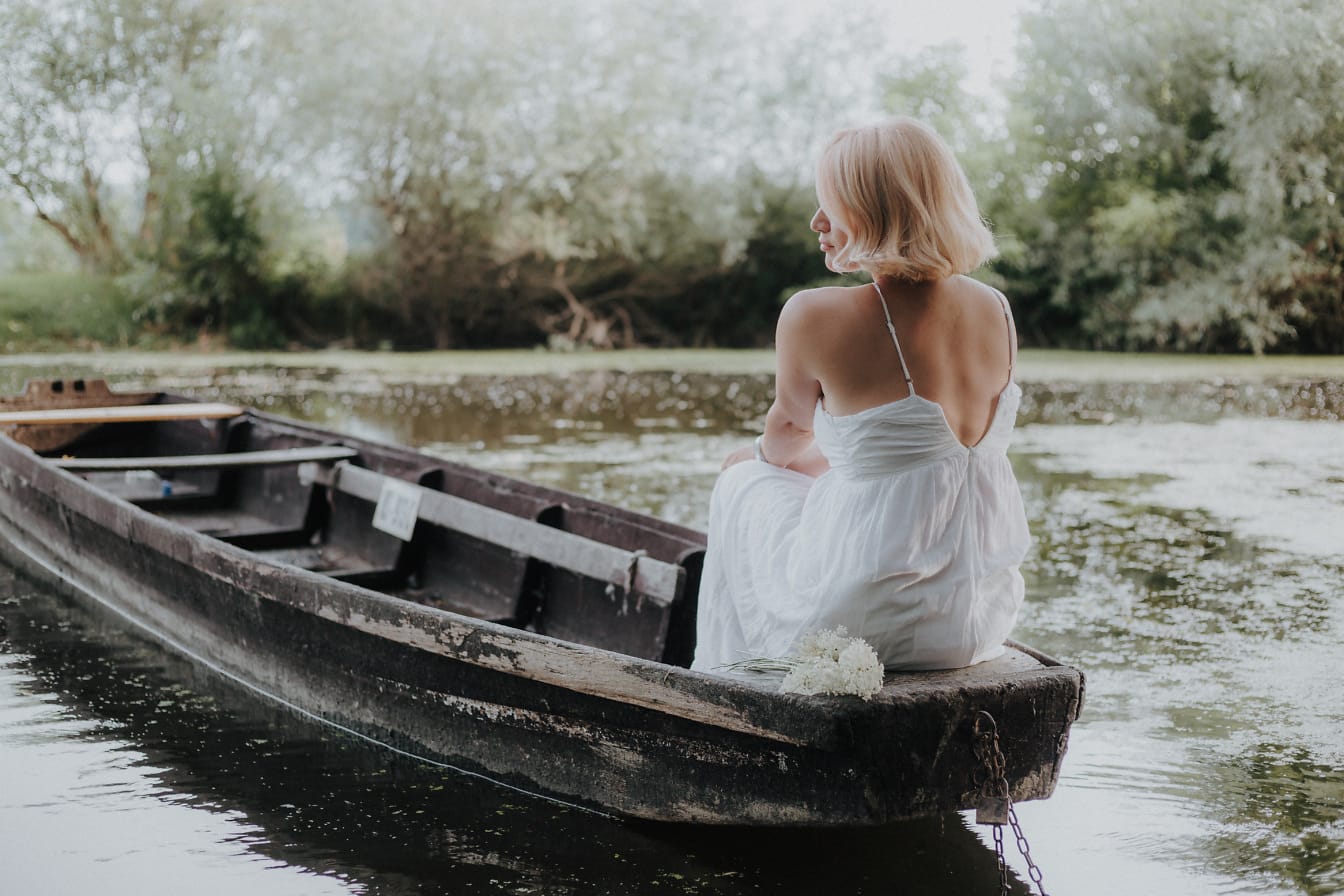 Wanita pirang cantik duduk di perahu kayu dengan gaun putih