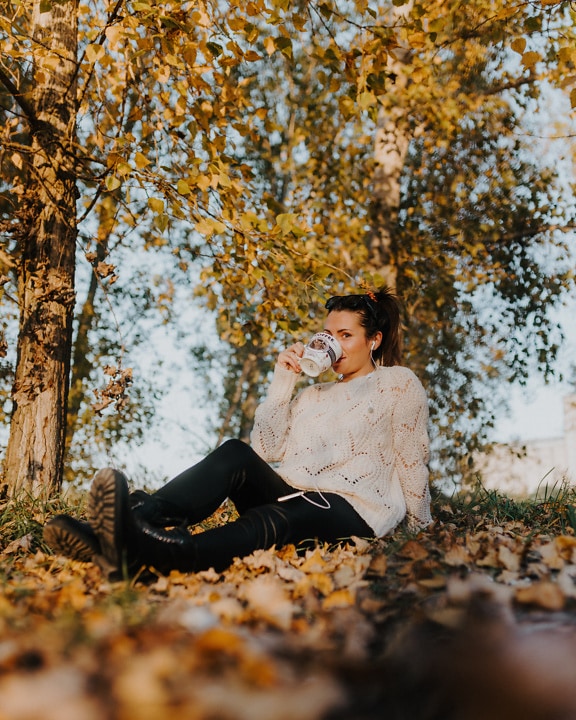 Nádherná brunetka sedí pod stromem a pije kávu z hrnku na kávu