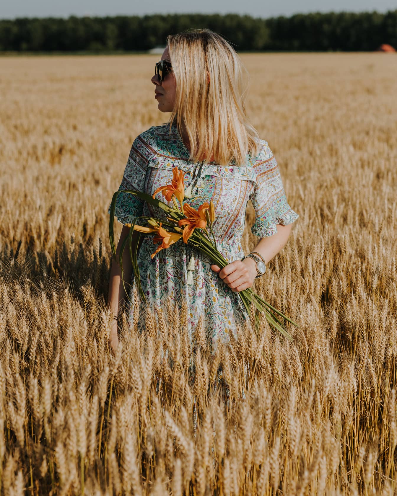 Femeie blondă de la țară stând în lan de grâu cu crini portocalii în mâini