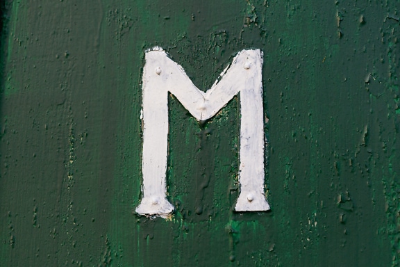 濃い緑色の塗装が剥がれた金属表面に白い文字(M)