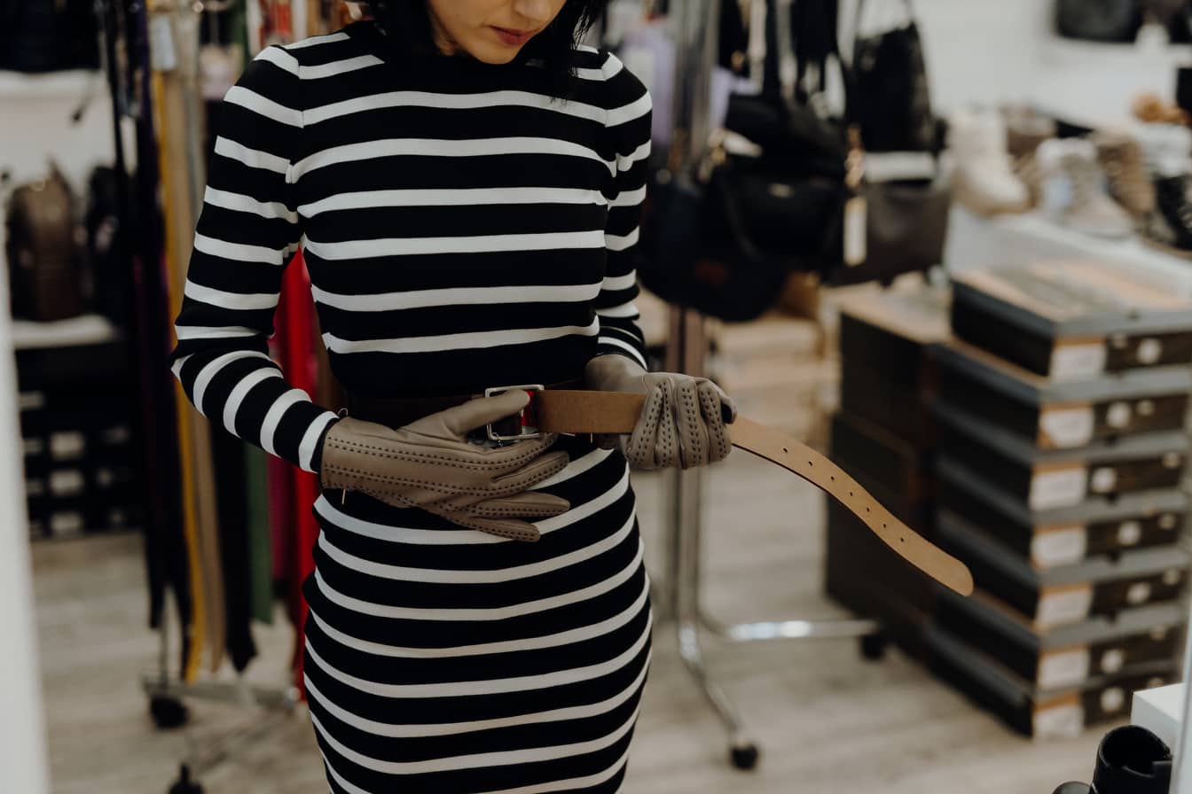 Женщина в полосатом платье и перчатках, держащая кожаный ремень в магазине