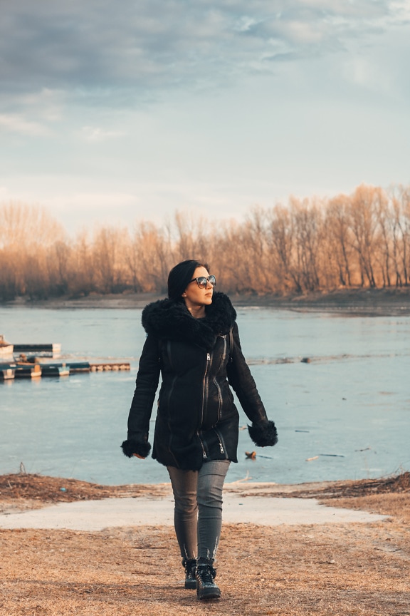 Wanita muda dengan mantel musim dingin dan kacamata hitam berjalan di pantai danau beku