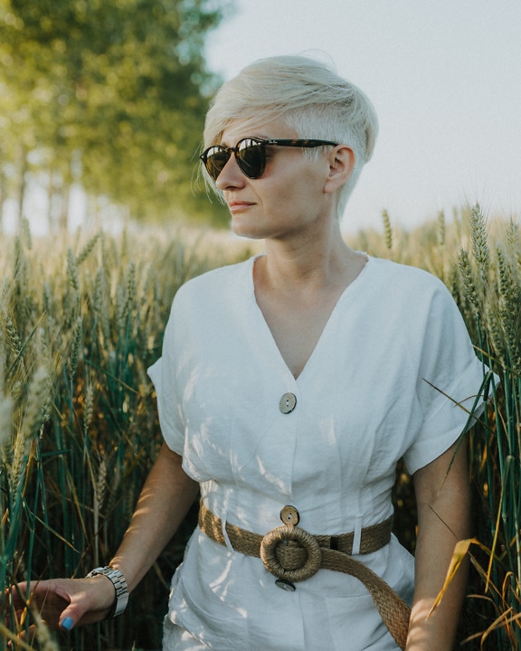 Dobře vypadající venkovská žena v pšeničném poli v bílých šatech a slunečních brýlích