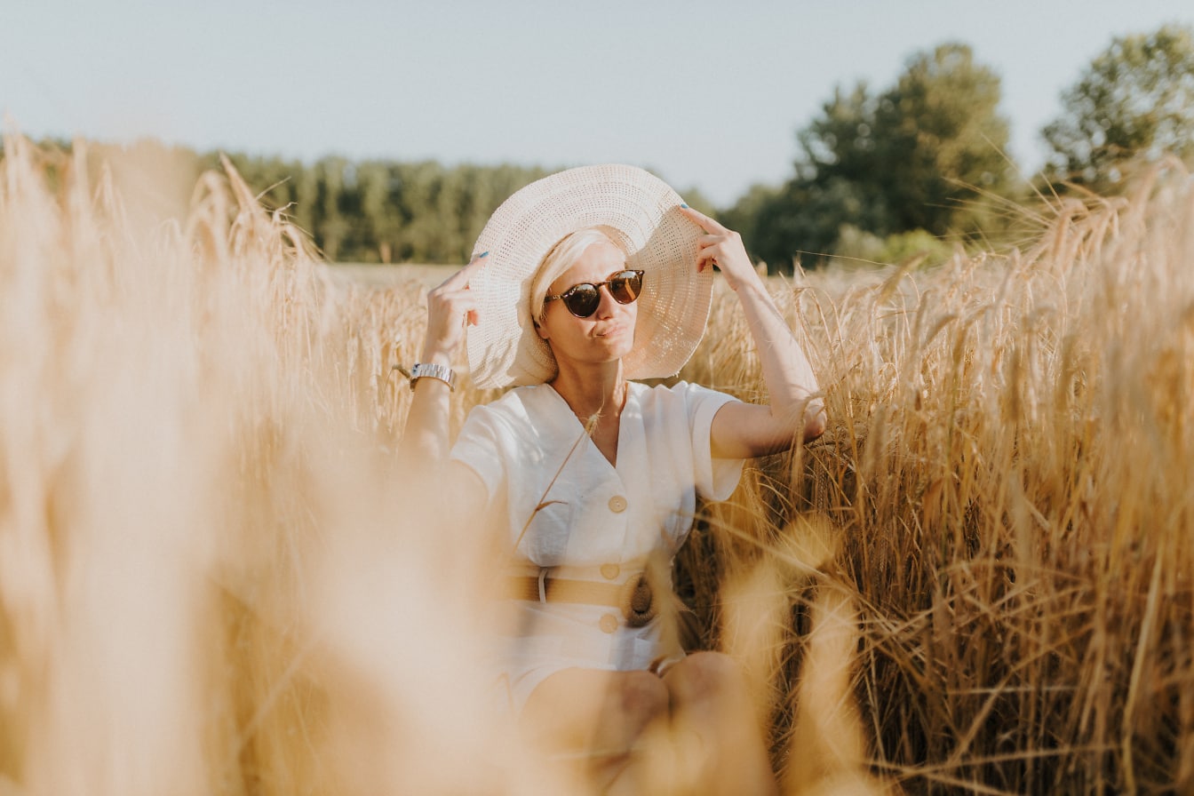 Pretty woman sitting in a field of wheat wearing elegant dress and straw hat at summer