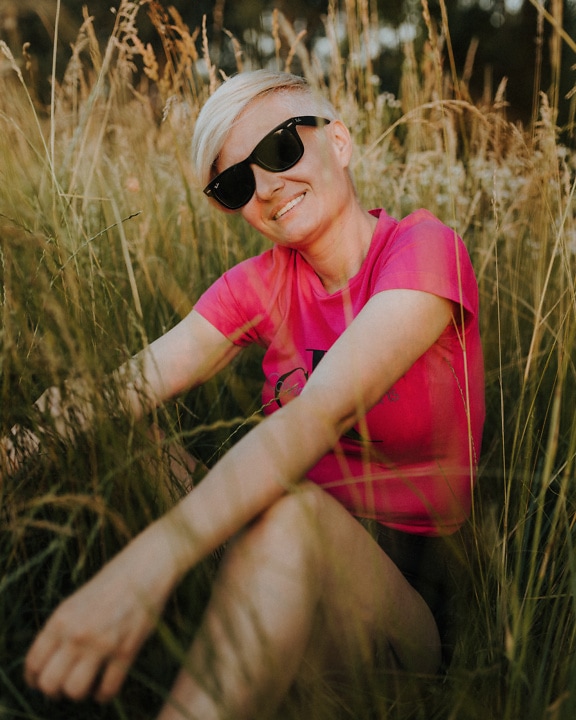 Ung kvinne med solbriller sittende i høyt gress og smilende