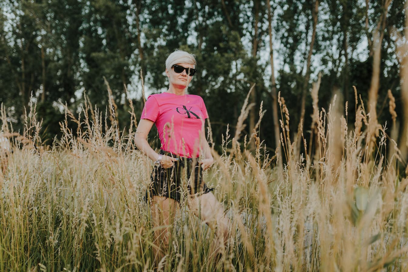 Rózsaszín inget és rövid nadrágot viselő nő büszkén pózol egy magas füves mezőn