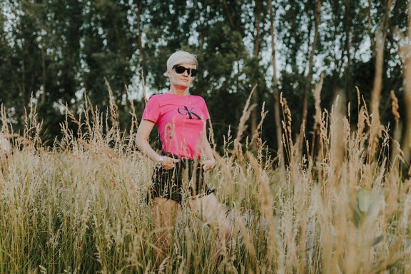 Žena v růžové košili a krátkých kalhotách hrdě pózuje na poli s vysokou trávou