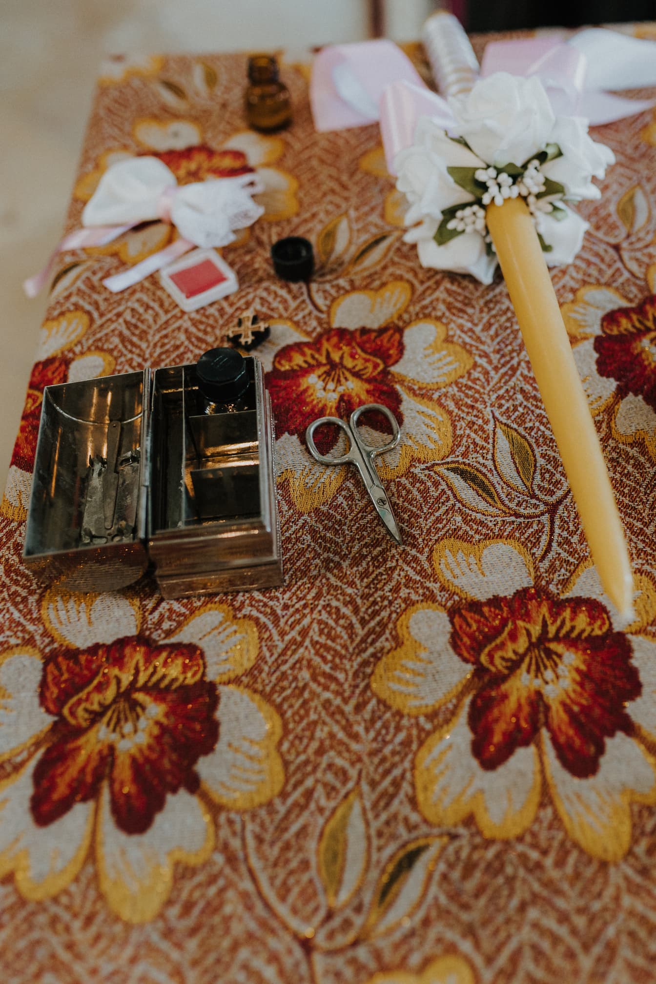 Метална кутия с ножица и свещ върху флорална покривка, подготвена за кръщелна церемония