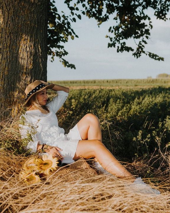 Maalaistyttö valkoisessa mekossa ja olkihatussa istumassa pellolla puun alla
