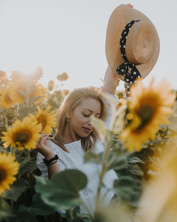 Вродлива весела молода жінка на полі соняшників із солом’яним капелюхом у руці