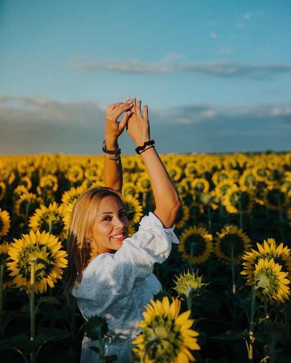 Người phụ nữ trẻ tóc vàng lộng lẫy vui vẻ trên cánh đồng hoa hướng dương với hai tay giơ lên không trung