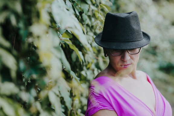 Dama elegante con sombrero negro y gafas posando junto al arbusto de hiedra