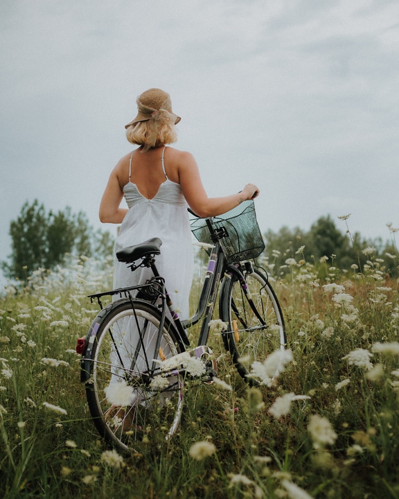 Maaseudun nuori nainen valkoisessa mekossa polkupyörän kanssa kukkaniityllä