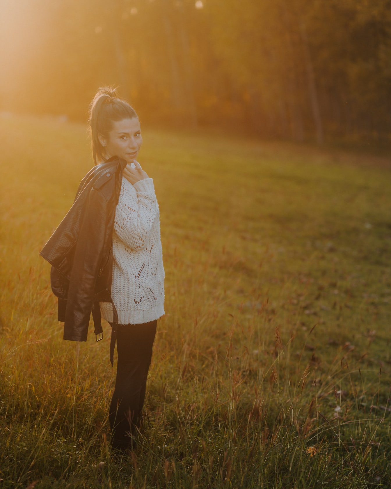 Женщина, стоящая в поле в кожаной куртке на фоне светящихся солнечных лучей