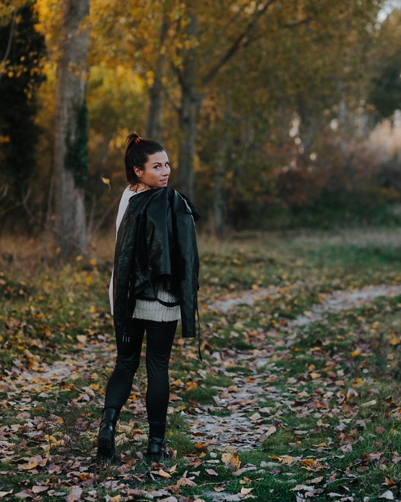 Woman standing on a forest path holding leather jacket in hand with forest in background