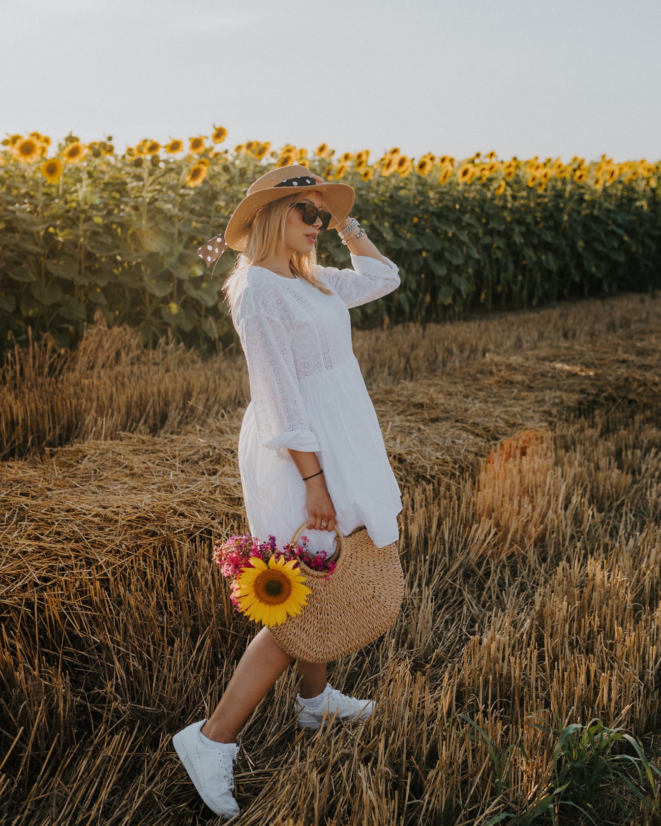 Donna nel campo di grano in un vestito bianco e cappello che tiene una borsa e fiori