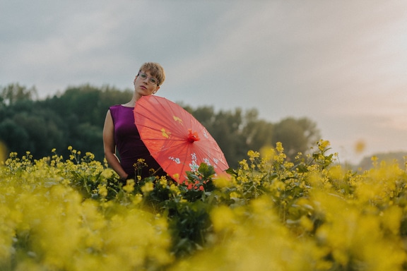 Femme glamour tenant un parapluie rouge dans un champ de fleurs de colza jaunes