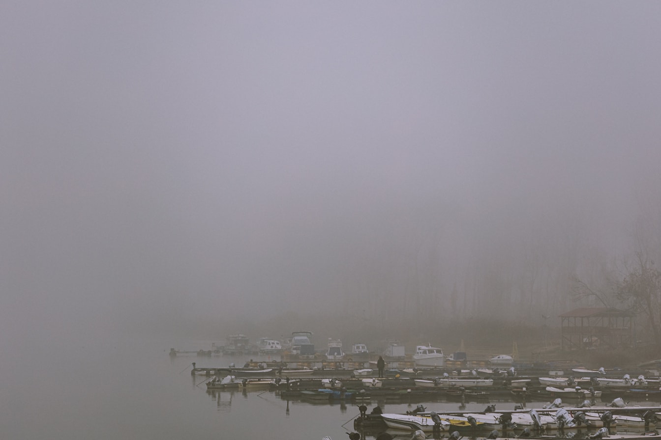 Båtar på en sjö i tät dimma med person som står på hamnen