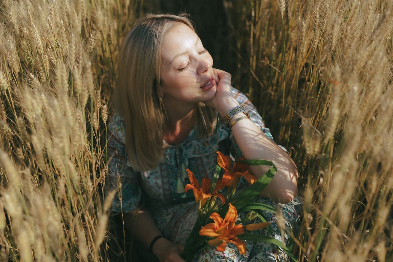 Pen blondine som sitter i en hveteåker med blomster i hendene og soler seg