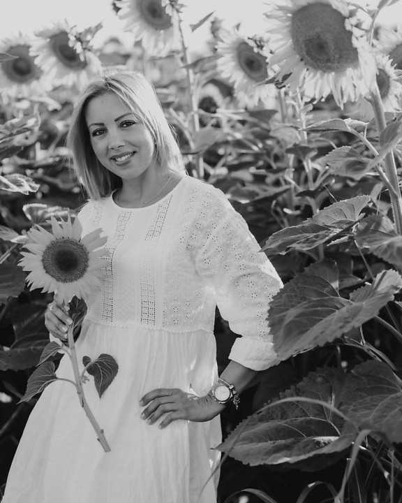 Tânără blondă zâmbitoare într-o rochie albă ținând o floarea-soarelui