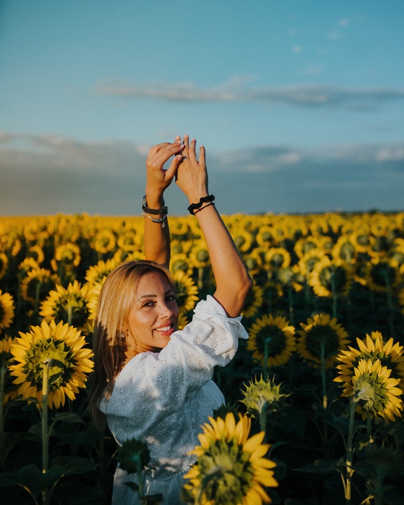 Underbar blond kvinna i ett fält av solrosor som håller hennes händer upp i luften