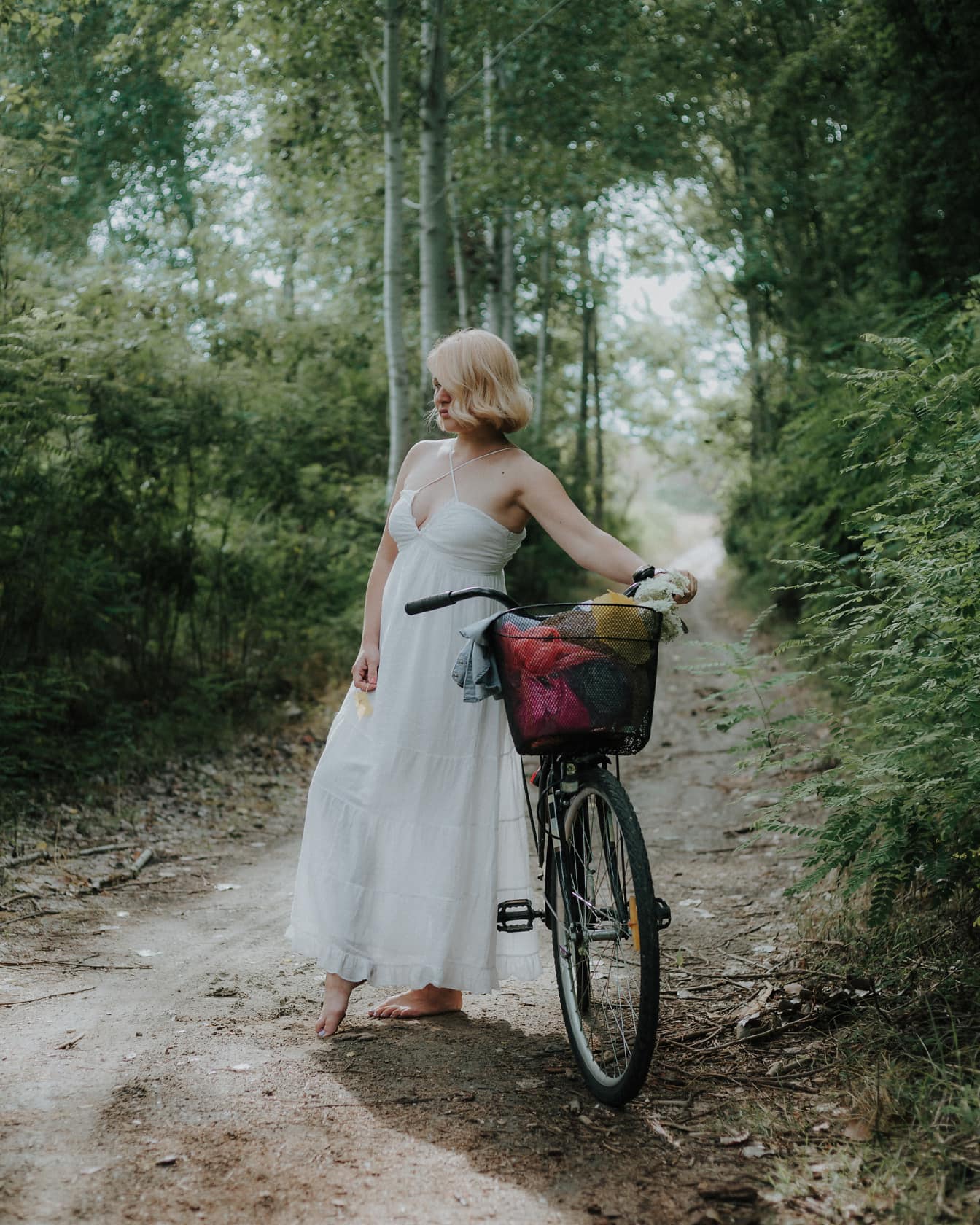 Signora bionda a piedi nudi in un vestito bianco con una bicicletta su un sentiero sterrato nella foresta