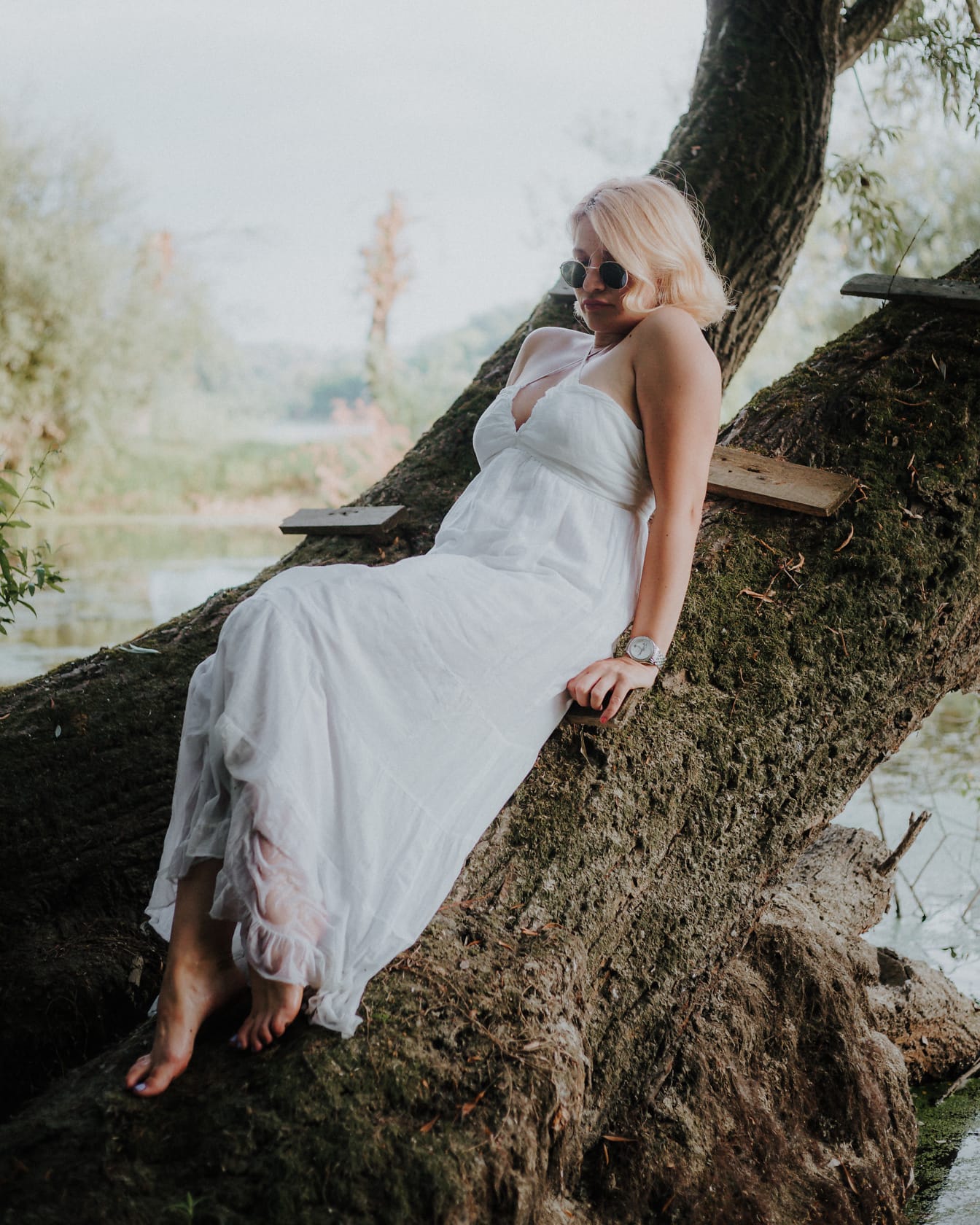 Wunderschöne barfüßige verführerische Frau in einem weißen Kleid auf einem Baum liegend