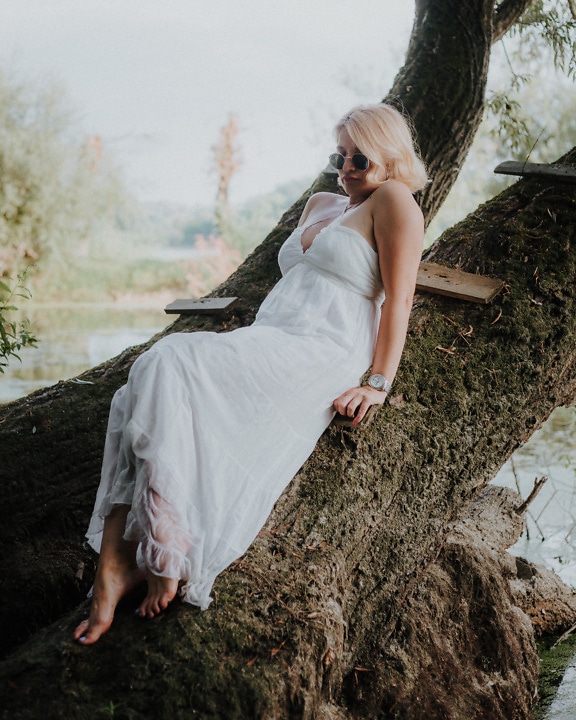 Gorgeous barefoot seductive woman in a white dress lying on a tree