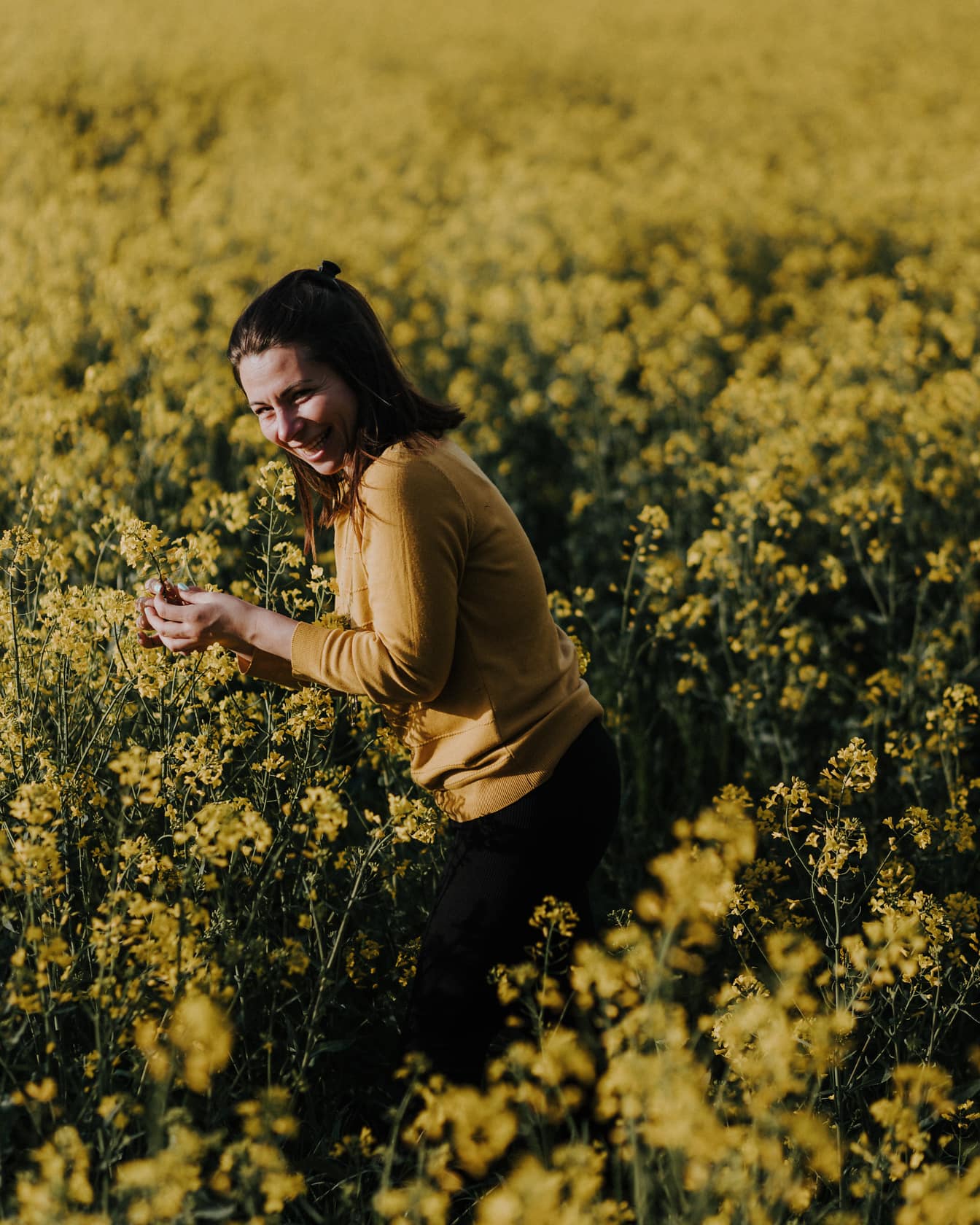 Femeie brunetă zâmbitoare stând într-un câmp de rapiță cu flori galbene