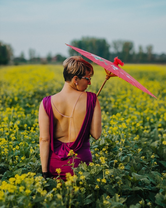 Mulher no vestido roxo sem costas segurando um guarda-chuva vermelho em um campo de flores amarelas