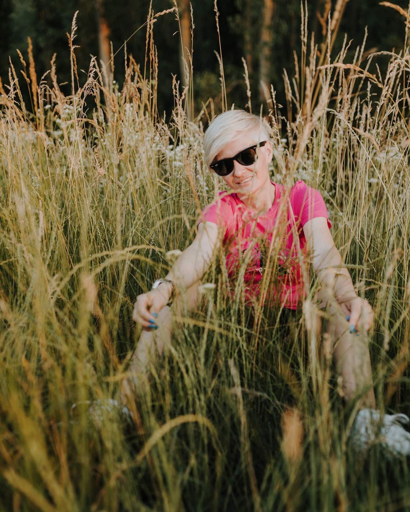 Usmievavá žena s krátkym blond účesom sediaca vo vysokej tráve
