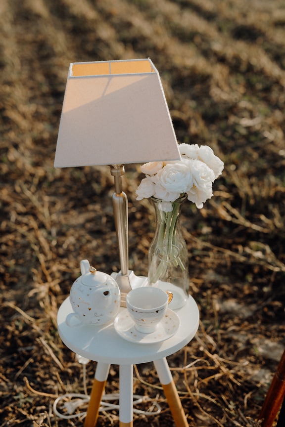 Stôl s modernou lampou, porcelánovou kanvicou a sklenenou vázou s kvetmi bielej ruže