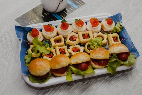 Поднос со свежими миниатюрными бутербродами, гамбургерами и другими закусками на столе