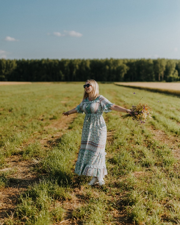 Glad kvinde på landet i en traditionel kjole, der holder blomster på en mark