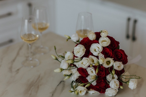 Kytice červených a bílých růží na mramorovém stole se skleničkami bílého vína v pozadí