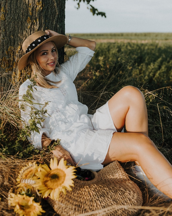 Cowgirl drăguță de la țară așezată pe un câmp în rochie albă și pălărie de paie